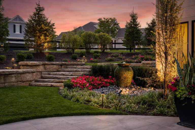 Landscaped retaining wall, stone steps, planting beds and a lit water feature at dusk