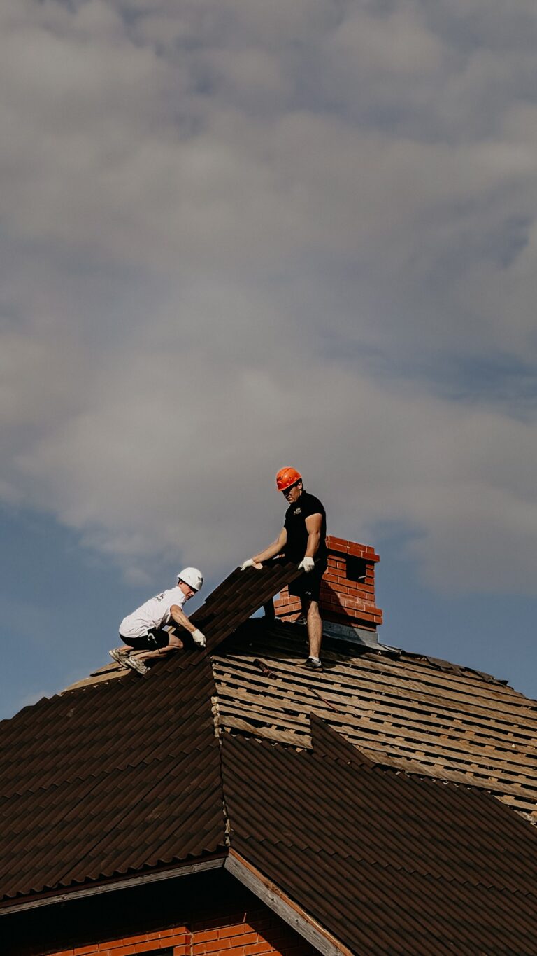 Two roofers in hard hats replacing tiles on a steep roof beside a brick chimney
